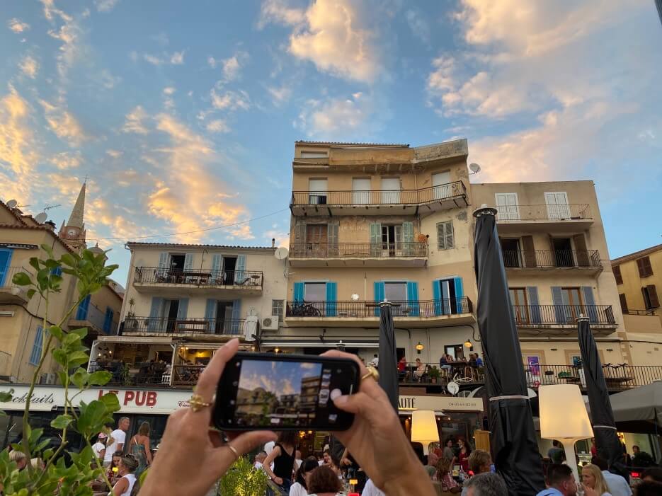 Photo of someone taking a photo with a phone of pretty village buildings and crowded cafes