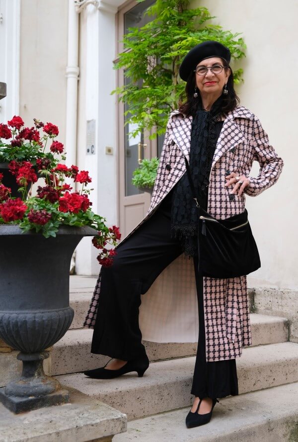 Adrian Leeds in black pants and long black and white coat in her Paris courtyard