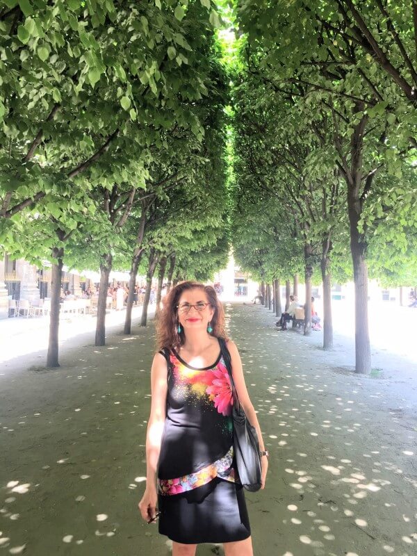 Adrian Leeds in a summer dress, standing on a tree lined pathway in a Paris park