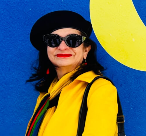 Adrian Leeds dressed in a bright yellow jacket and signature black beret in front of a blue background