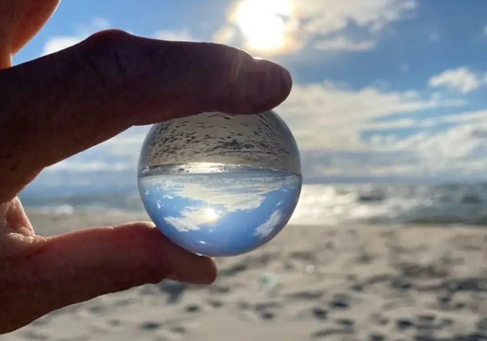 Person on the beach holding a small crystal ball in their fingers looking at the sky and water